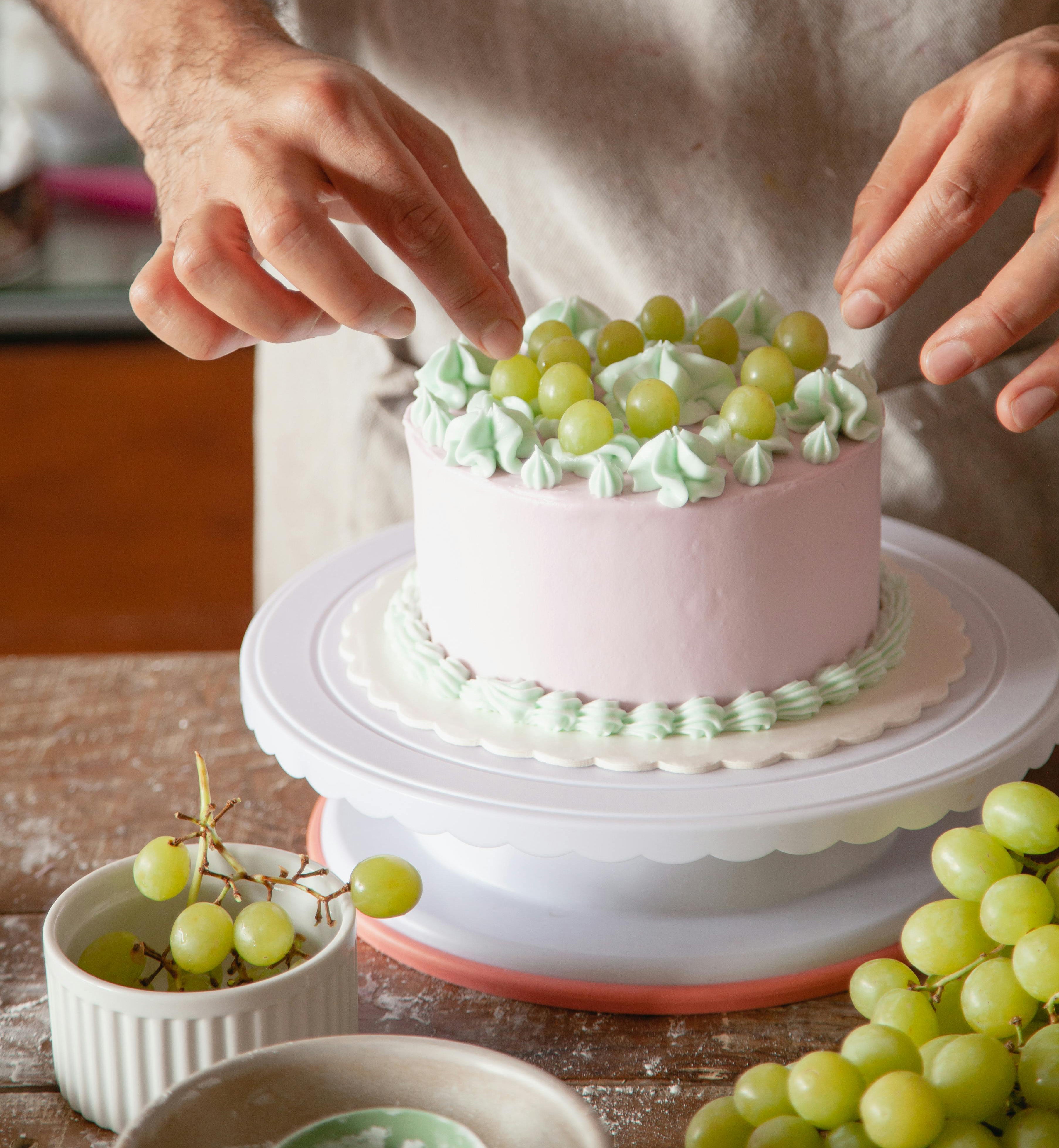 Person decorating a pink cake with green grapes on a wooden table.